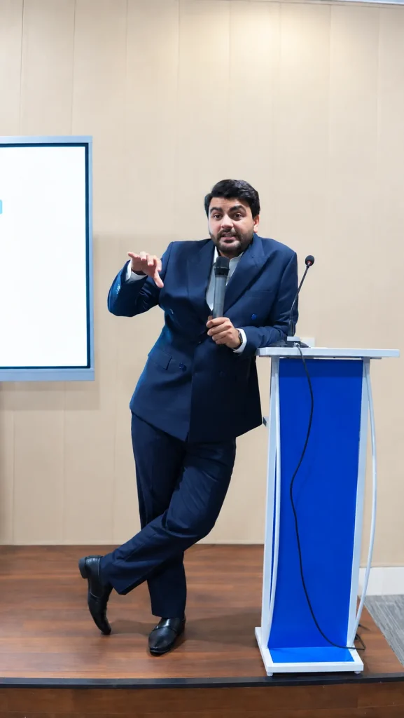 Umar Hameed on stage leaning on a podium, holding a microphone and pointing to the audience