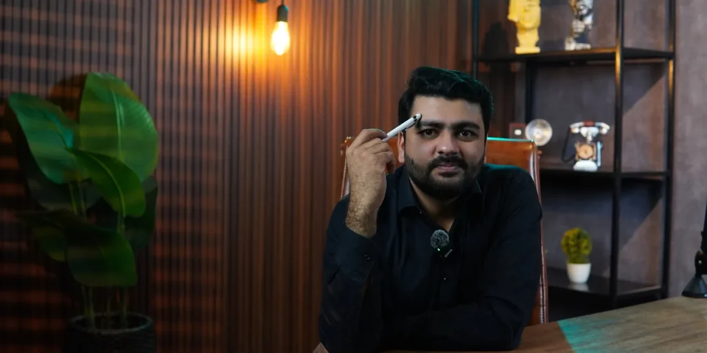 Umar Hameed in a black shirt seated at a dark wood desk, holding his glasses