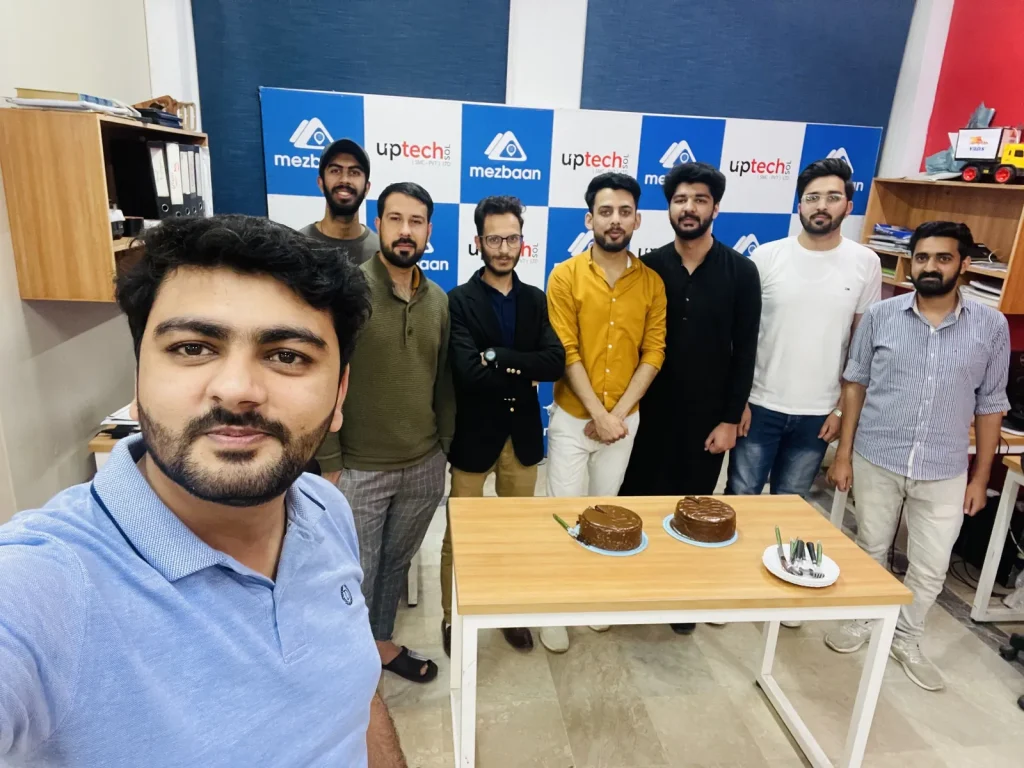 Umar Hameed taking a selfie with eight people standing behind a table with two cakes