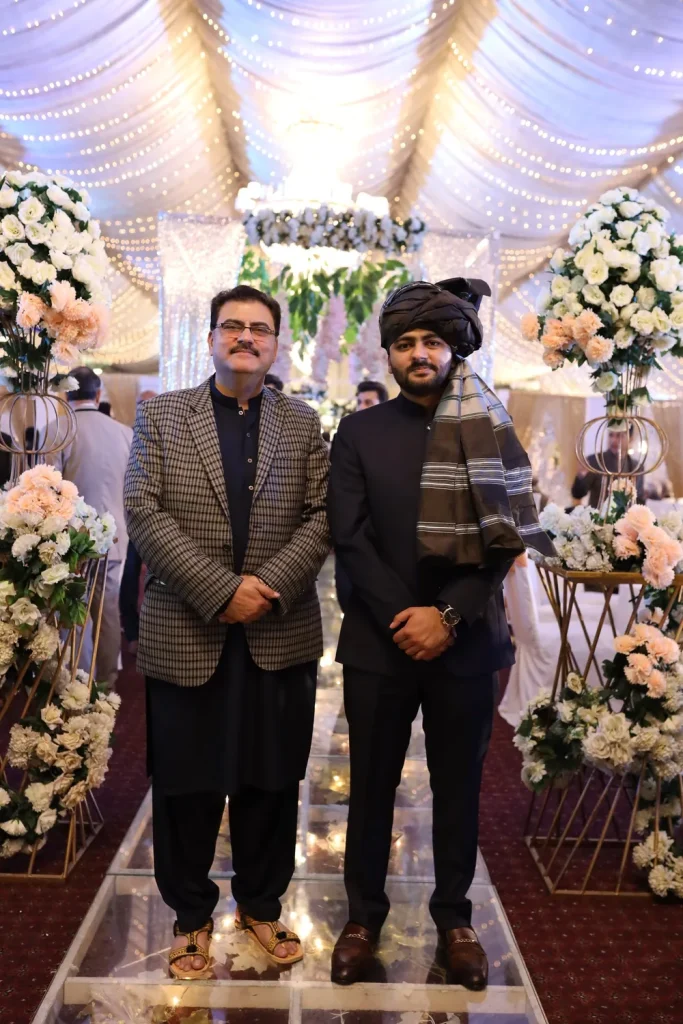 Umar Hameed standing beside his father, who wears glasses and a checked blazer, with white and peach flowers behind them