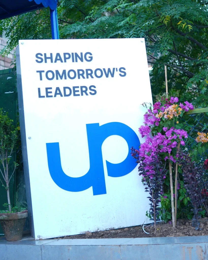 Uptech Institute sign reading Shaping Tomorrow's Leaders with a large blue UP logo, beside a flowering bush
