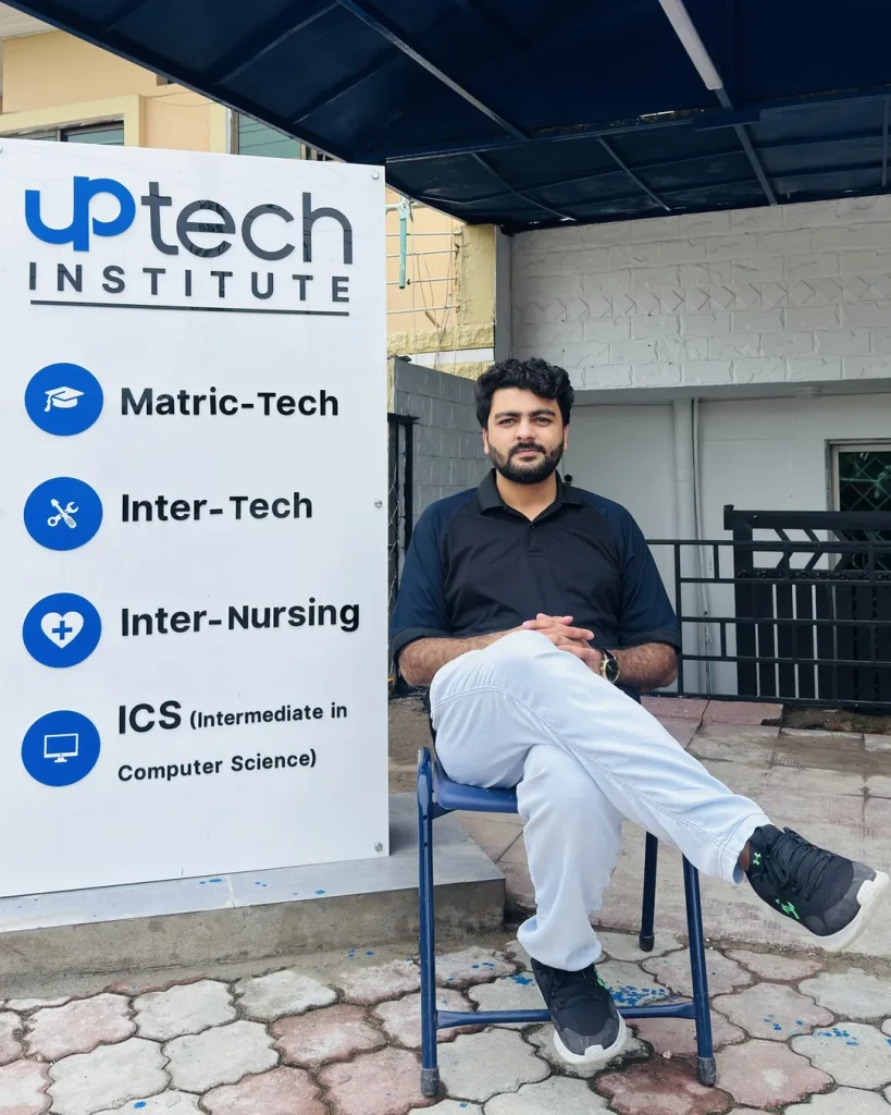 Umar Hameed sitting on a chair beside the Uptech Institute signboard listing Matric-Tech, Inter-Tech, Inter-Nursing and ICS