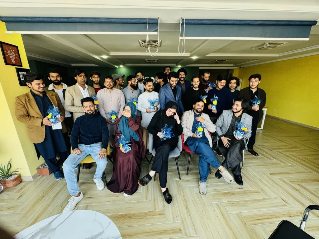 Umar Hameed with a large group of about thirty colleagues posing in a yellow-walled room, many holding small blue gift bags